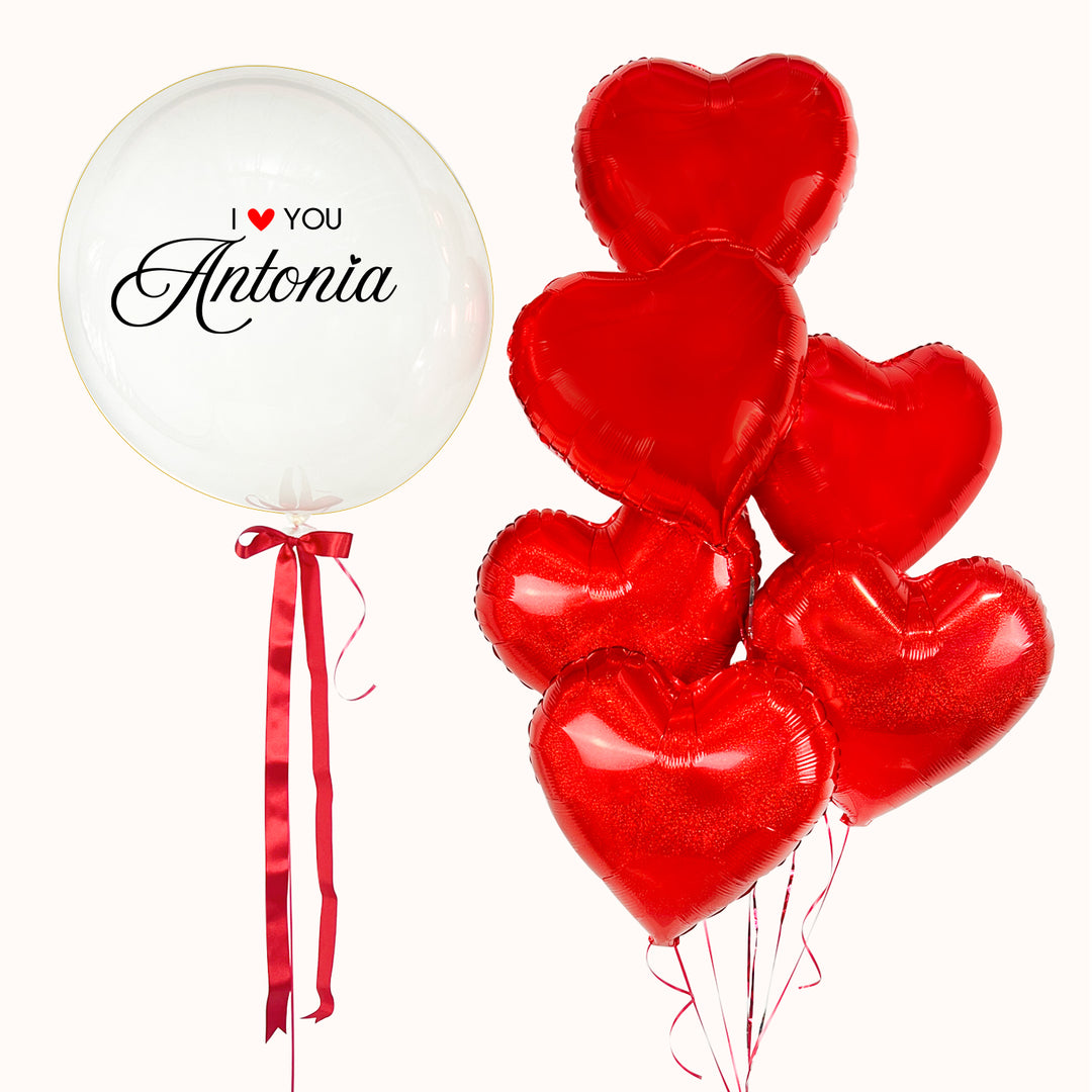 Arreglo de globos compuesto por 1 globo burbuja transparente personalizado, acompañado de 6 globos metálicos en forma de corazón color rojo. Para sorprender en San Valentín o ocasiones especiales. Regalos de Aniversarios Románticos.