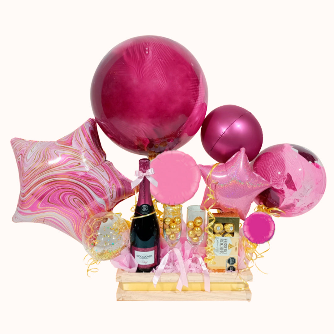 Gift box en tonos rosados y fucsia, que incluye bebida Riccadonna Rubí, dos copas rellenas con chocolates circulares y una caja de bombones Ferrero Rocher de 8 unidades, decorado con globos burbuja y globos metálicos circulares y de estrellas en diversos tamaños. Detalle para regalo de San Valentín, aniversario, cumpleaños o en ocasión especial. Regalos personalizables.