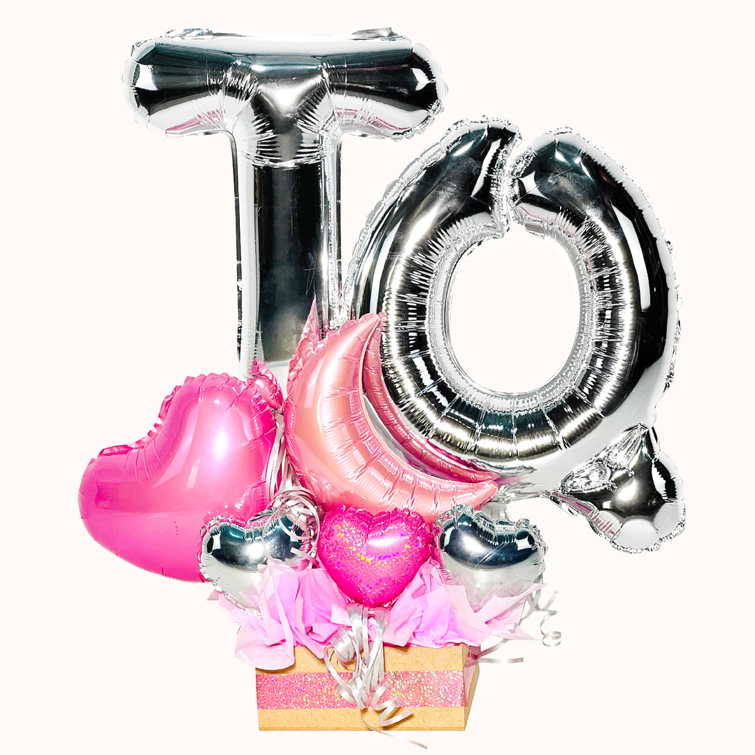 Gift box de madera con globos de letras metálicas 'TQ' en color plata y arreglo de mini globos en tonos rosado y plateado para declarar amor.