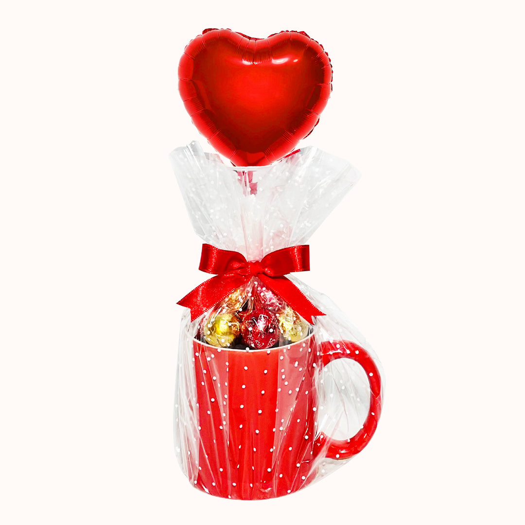 Taza de cerámica rellena con chocolates circulares, envuelta en papel celofán y acompañada de un globo metálico en forma de corazón personalizado con tu logo. Ideal para sorprender a clientes y colaboradores. 